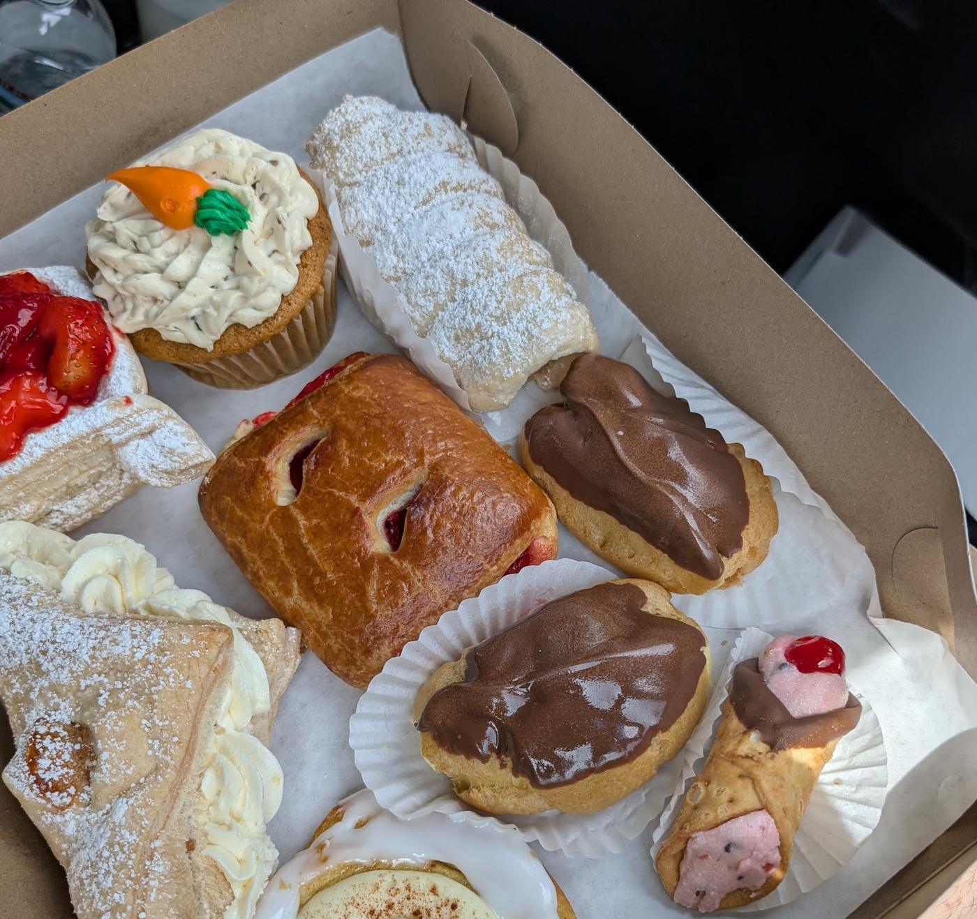 A bakery box packed at the counter: a carrot cupcake, a strawberry napoleon, a cannoli, chocolate custard puffs and a custard danish